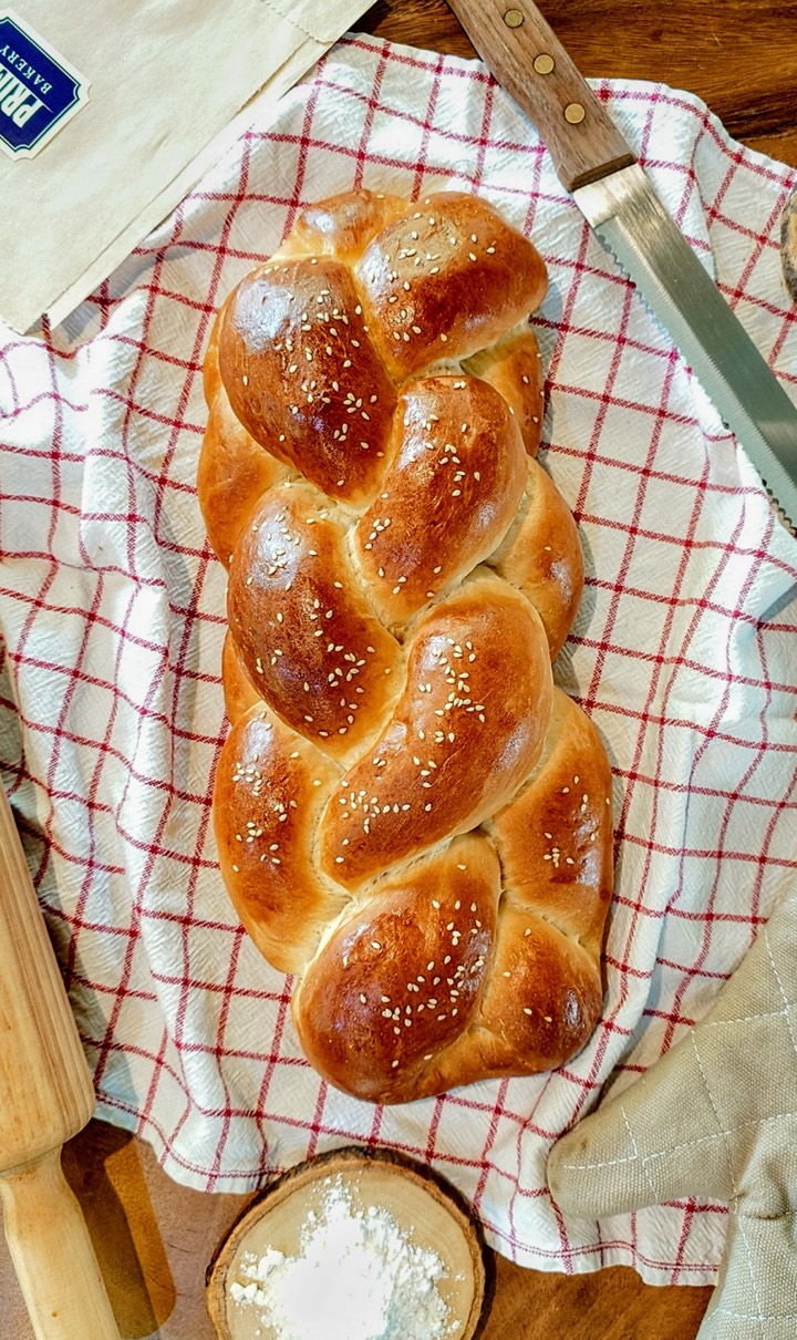 A golden braided challah loaf with sesame seeds on a red check cloth