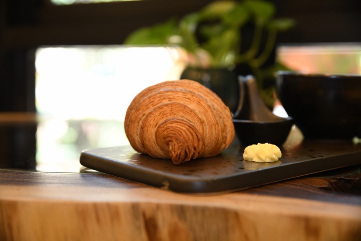 A golden, flaky croissant on a dark serving board with a swirl of butter, coffee cups and a plant softly in the background